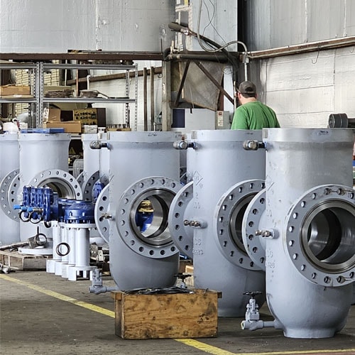 Row of large grey industrial valve bodies in a workshop with a worker in the background